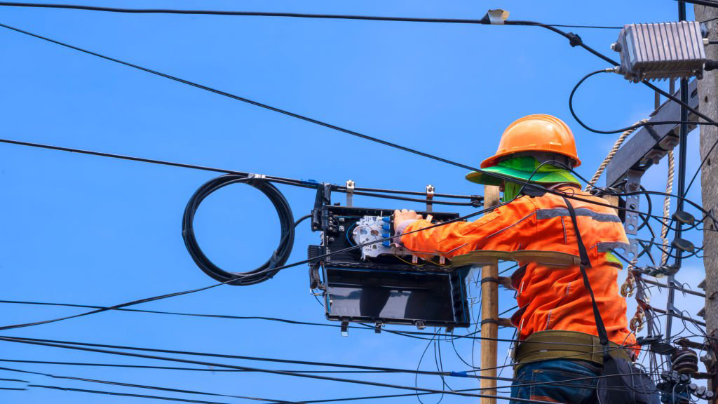 Rear view of technician on wooden ladder is working to install fiber optic and splitter box on electric pole against blue sky background