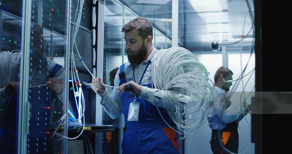 Medium shot of a male technicians working in a data center carrying cable down the corridor amongst rows of server racks