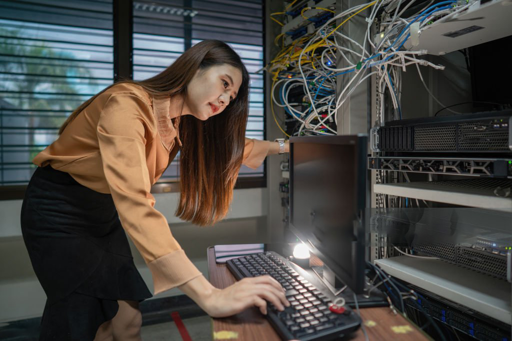 A female programmer is working in a server room. The girl is standing next to the computer racks of the data center. The administrator looks at the monitor with network settings.